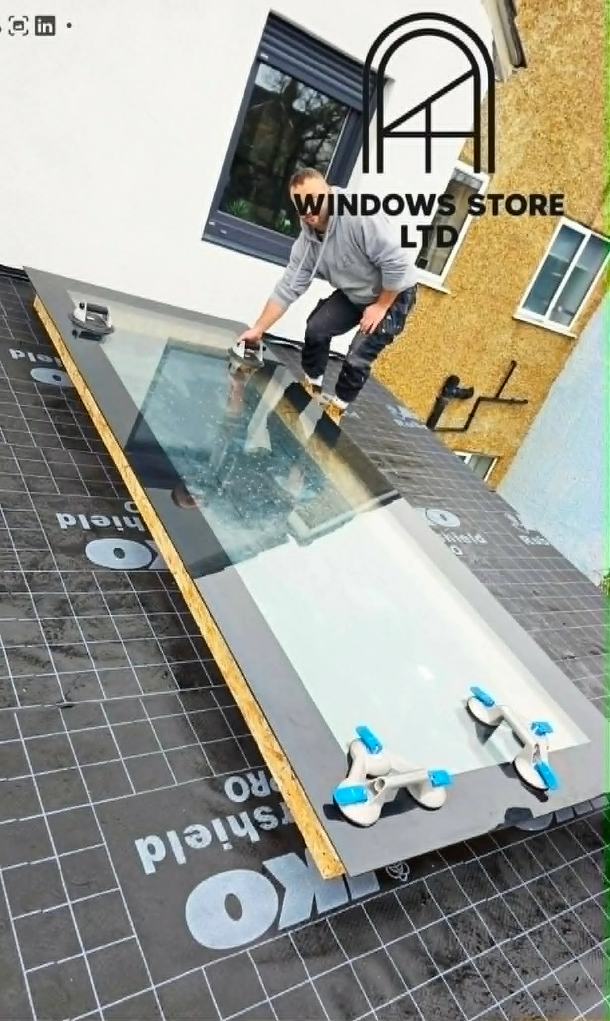 A large glass rooflight panel being lowered onto a flat extension roof, seen from above, with a fitter guiding it on suction handles — Windows Store Ltd, London